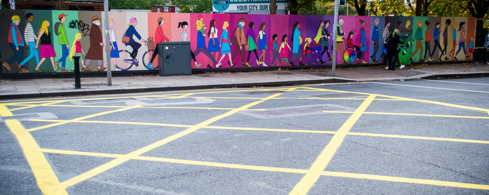 Kreuzung mit Wandbild im Hintergrund, auf denen diverse Menschen dargestellt sind 